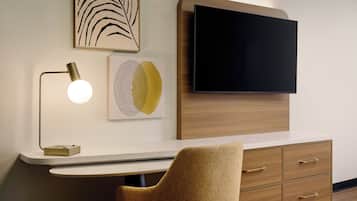Una habitación moderna con una televisión de pantalla plana montada en un panel de madera, un escritorio con lámpara, una silla y una obra de arte en la pared.