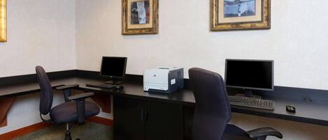 A hotel office with two desks, each with a computer monitor, keyboard, and mouse. There are two framed pictures on the wall.