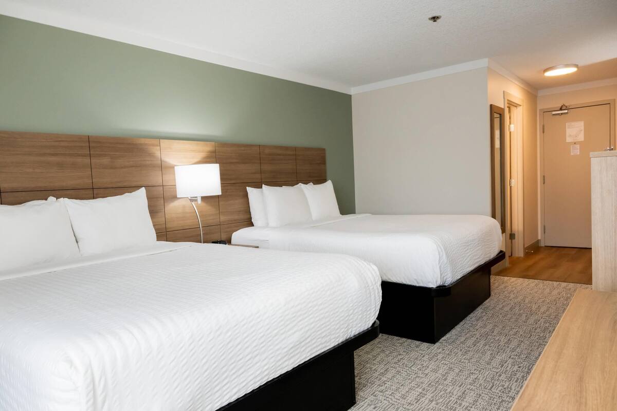 Habitación de hotel con dos camas, cabecero de madera, mesita de noche con lámpara y una puerta a la derecha.