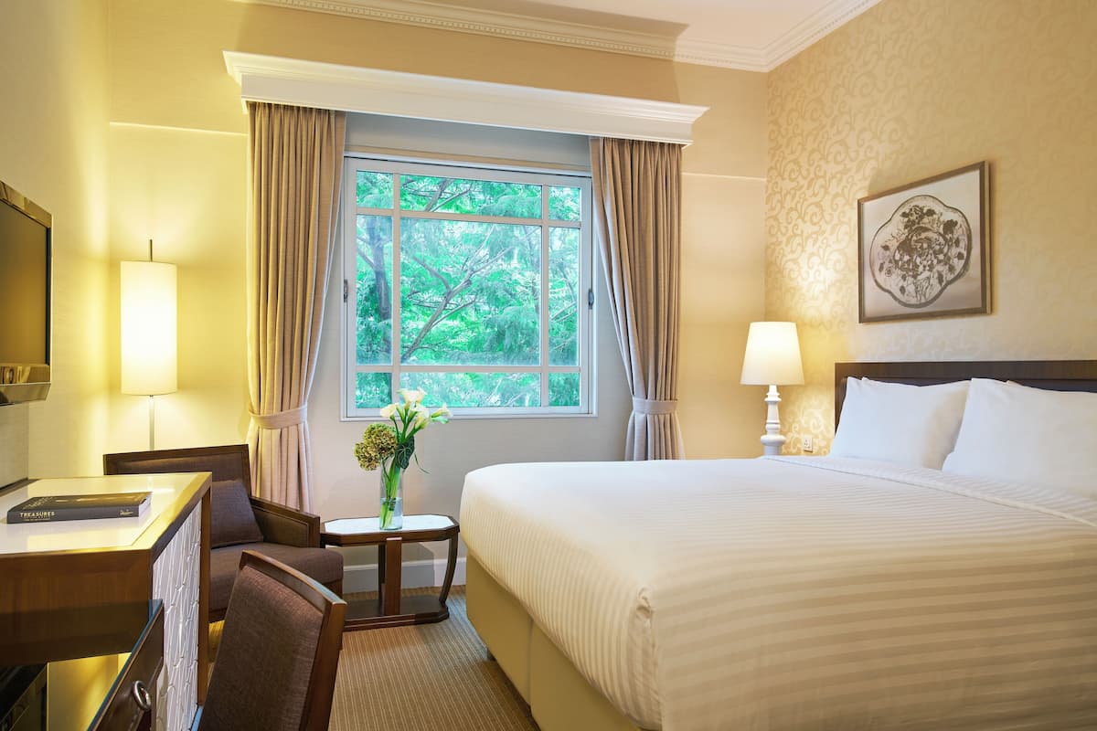 A hotel room with a large bed, a desk with a lamp, a chair, a small table with a vase of flowers, and a window with curtains.