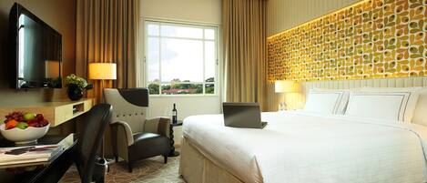 A hotel room with a large bed, a desk with a laptop, a TV, and a view of the city.