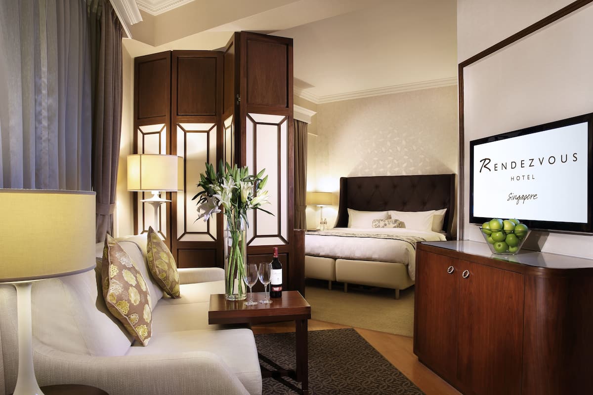 A hotel room with a bed, a sitting area with a sofa and armchair, a wooden cabinet, a table with a vase and wine glasses, and a framed sign on the wall.