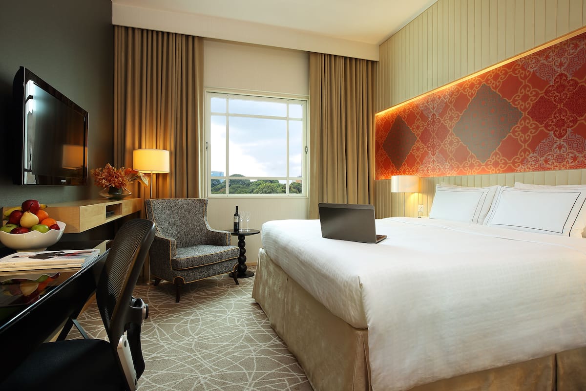 A hotel room with a large bed, a desk with a laptop, a chair, a TV, and a window with a view of greenery.