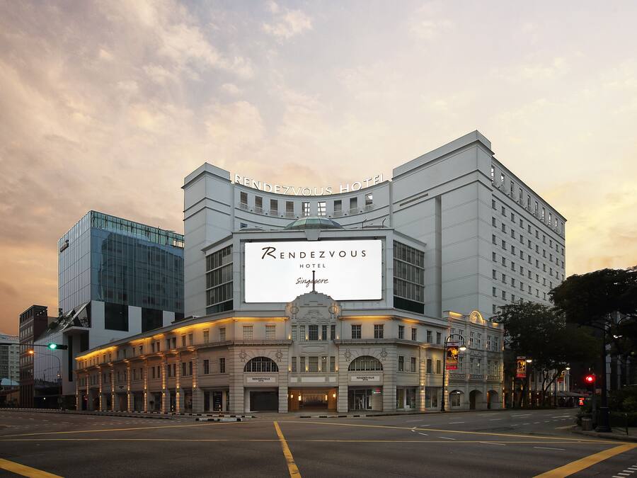 The exterior of the Rendezvous Hotel in Singapore, featuring a prominent sign, classical architecture, and a modern glass building adjacent to it.