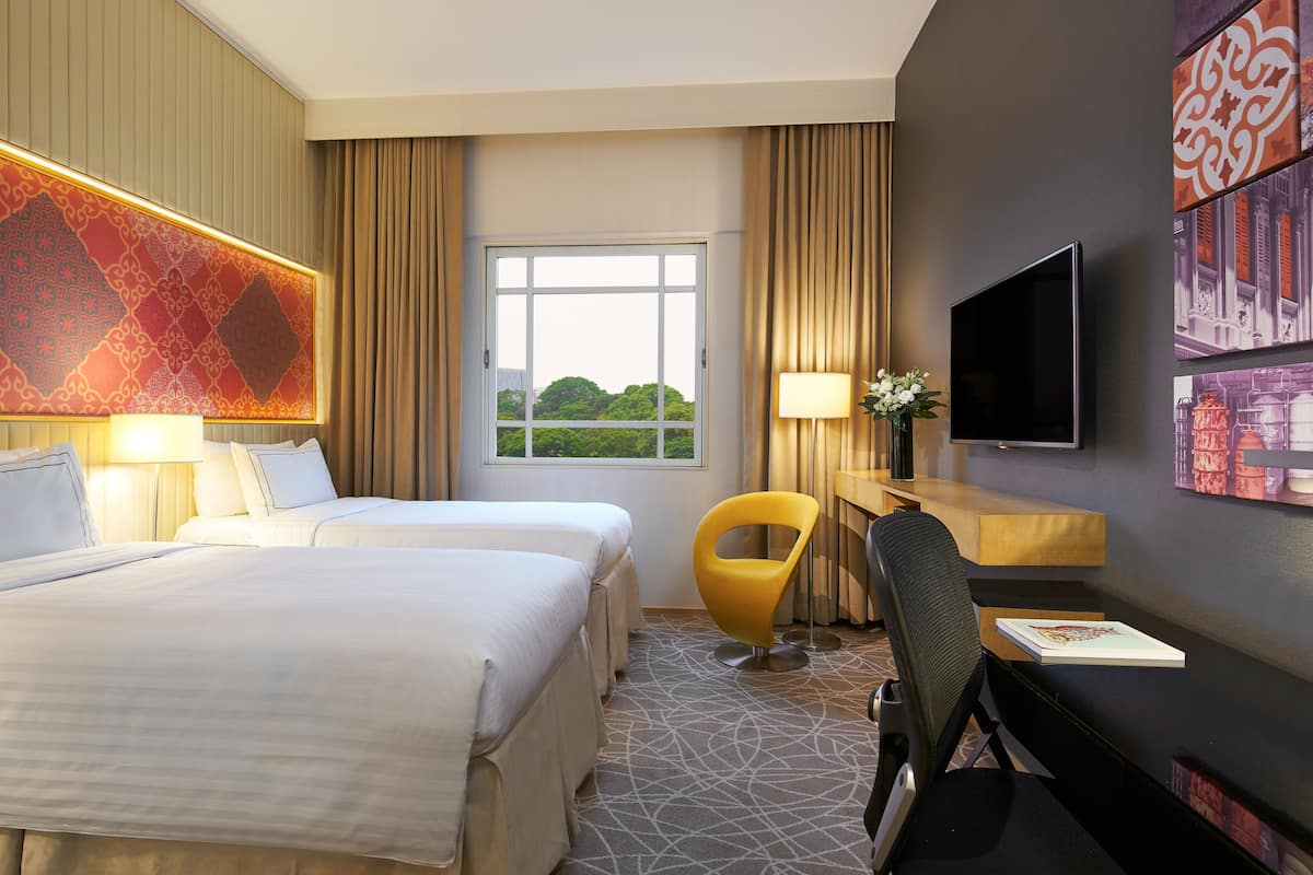 A hotel room with two beds, a desk, a chair, a TV, and a window with a view of greenery.