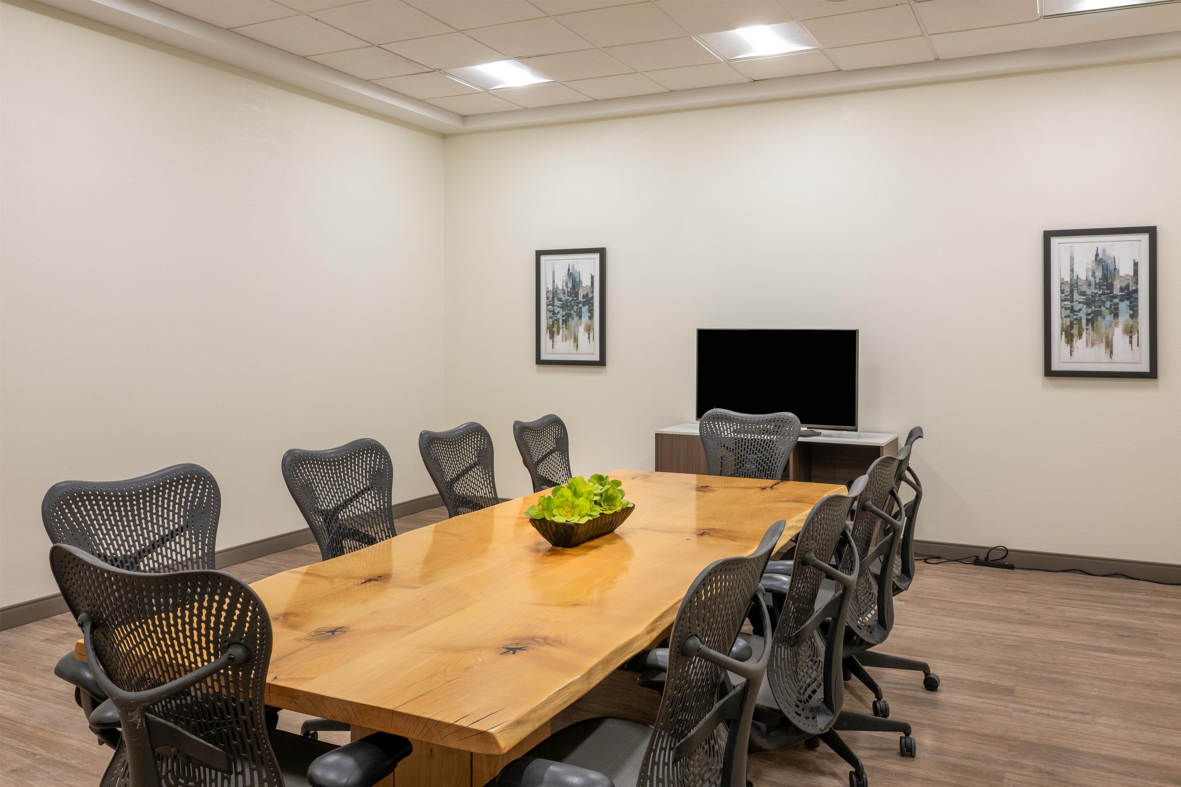 A conference room with a wooden table, black mesh chairs, a bowl of green plants, and two framed pictures on the walls.