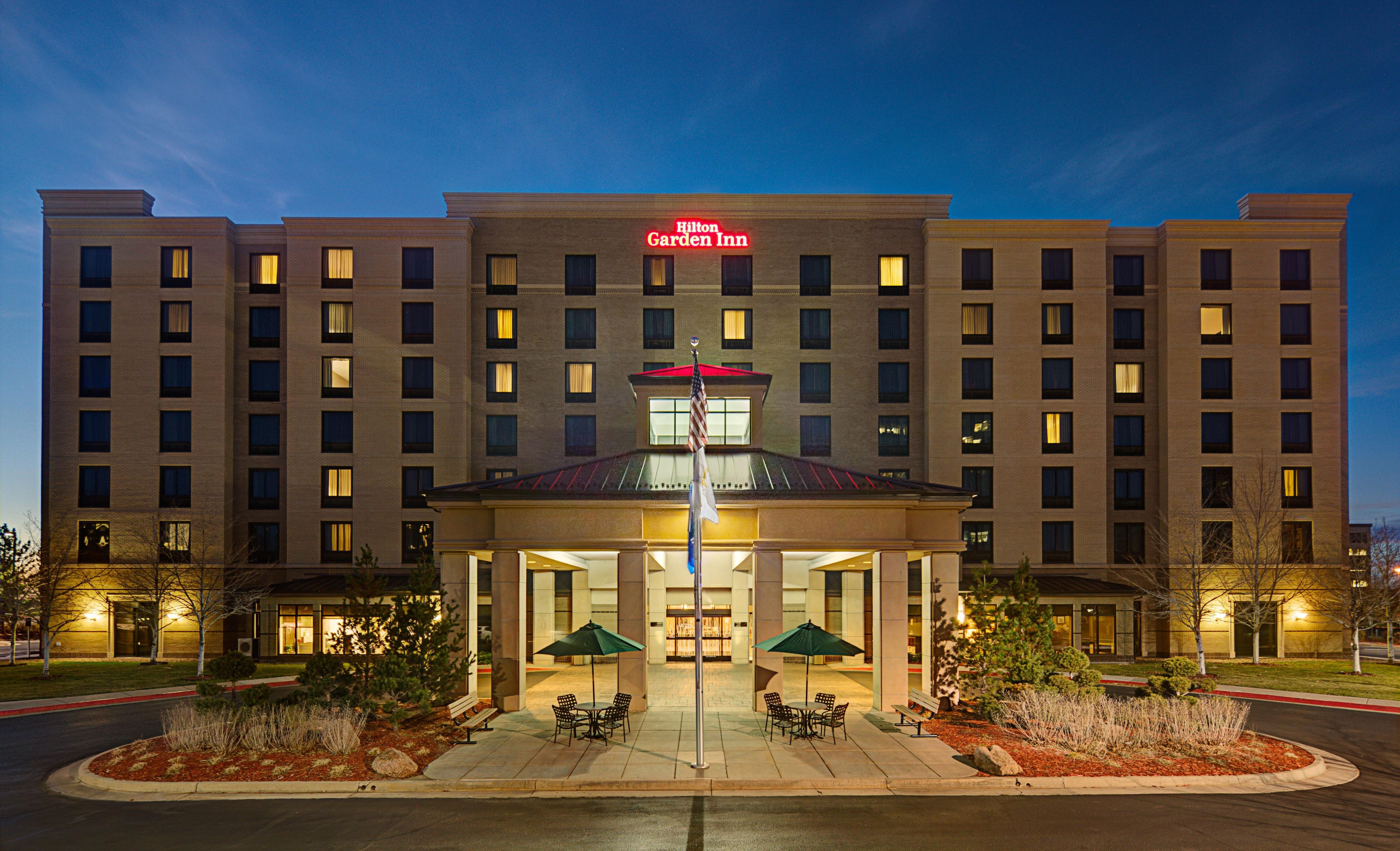 Un hotel a più piani con un cartello ben visibile con la scritta 'Hilton Garden Inn', un ingresso coperto con posti a sedere e un'asta sventolante con la bandiera americana.