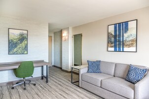 A modern hotel room with a desk, green office chair, grey sofa with patterned cushions, and two framed landscape paintings.
