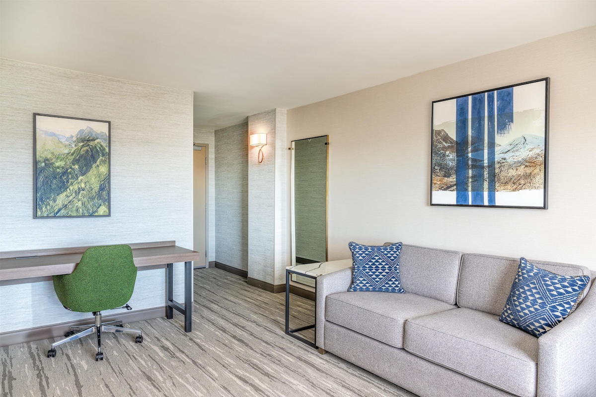 A modern hotel room with a desk, green office chair, grey sofa with patterned cushions, and two framed landscape paintings.
