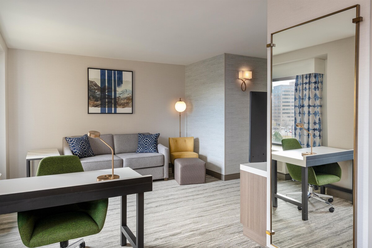 A hotel room with a desk, a sofa, a small ottoman, a green chair, a mirror, and a wall-mounted lamp.