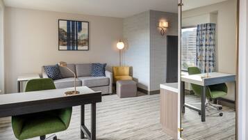 A hotel room with a desk, a sofa, a small ottoman, a green chair, a mirror, and a wall-mounted lamp.