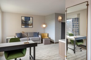 Ein Hotelzimmer mit Schreibtisch, einer Sitzecke, einem kleinen Hocker, einem grünen Sessel, einem Spiegel und einer Wandlampe.