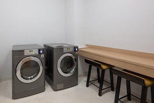 Un área de lavandería con dos lavadoras de carga frontal y una mesa de madera con taburetes negros.