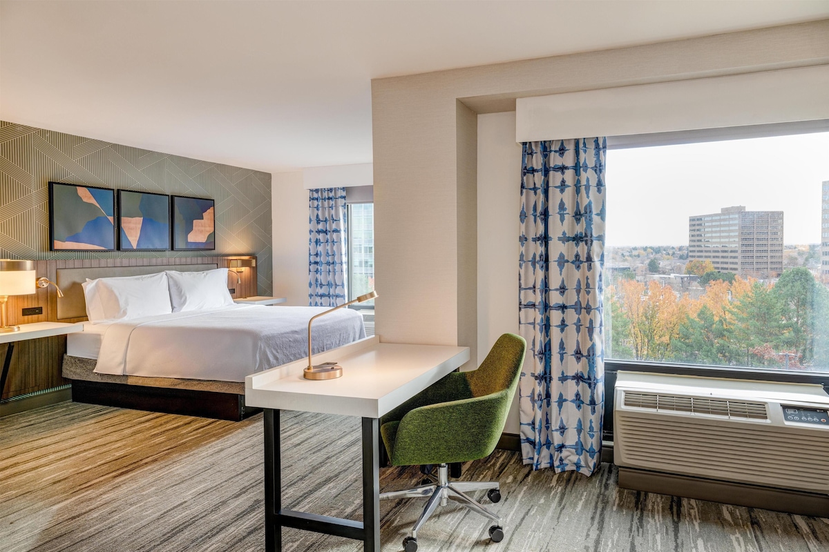 A hotel room with a bed, desk, chair, air conditioner, and a city view through the window.