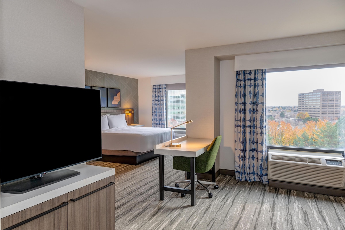 Quarto de hotel com cama, escrivaninha, cadeira, televisão e amplas janelas com vista para a cidade.