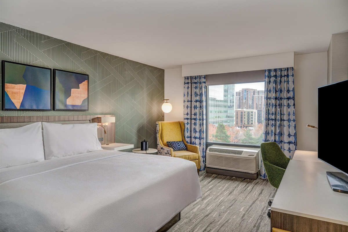 Une chambre d’hôtel avec un grand lit, un bureau, une chaise, une télévision, une fenêtre avec des rideaux et des œuvres d’art accrochées au mur.