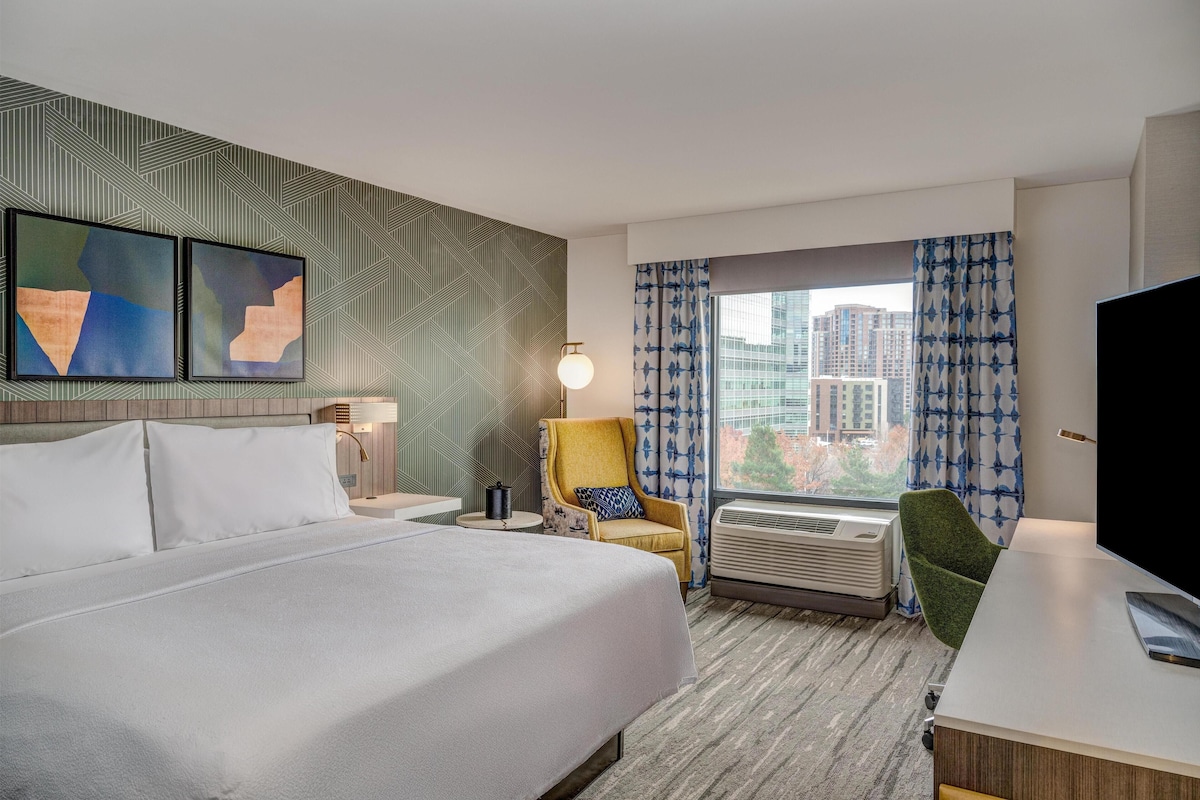 A hotel room with a large bed, a desk, a chair, a TV, a window with curtains, and artwork on the wall.