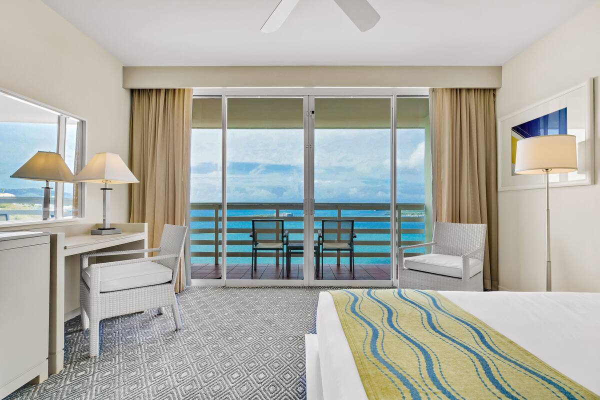 Habitación de hotel con cama, escritorio, dos sillas, un ventanal con vista al océano y un ventilador de techo.