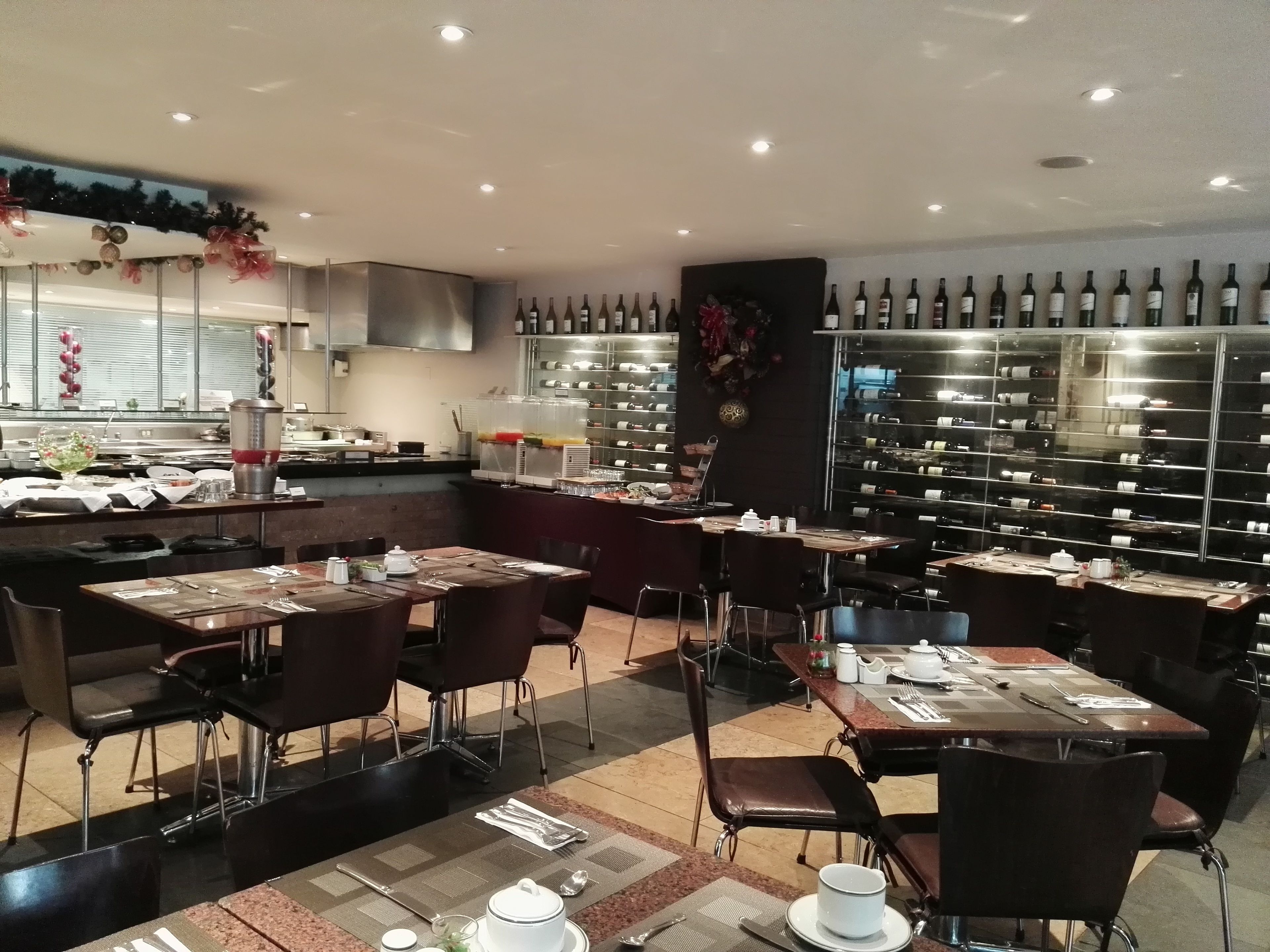 En restaurant med flere borde og stole, et køkkenområde og vinflasker på hylder.