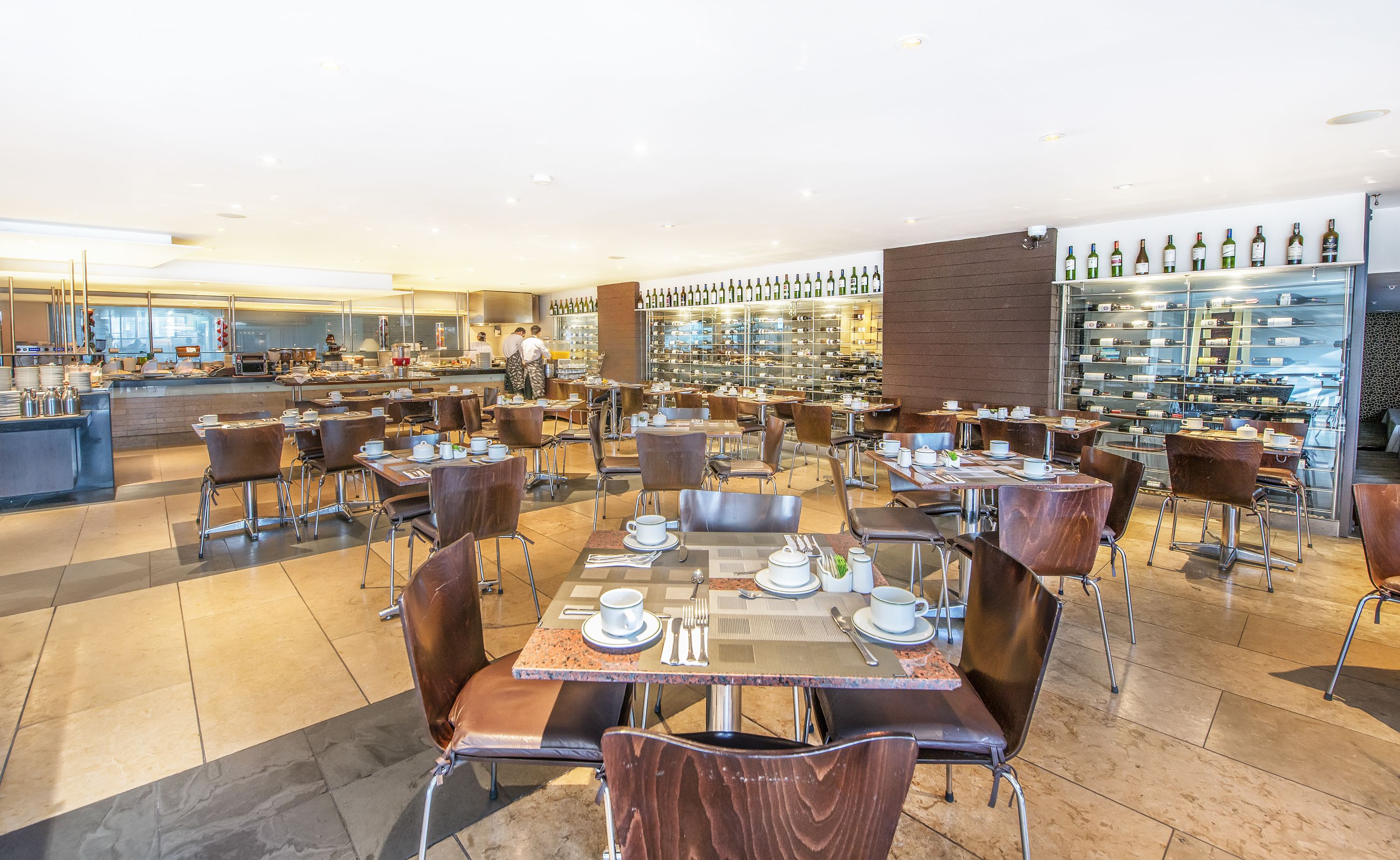 Un grand espace dédié à la salle à manger, avec plusieurs tables et chaises, une cuisine à l’arrière-plan et un présentoir à vin sur le côté droit.