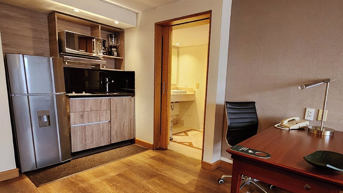 A compact kitchen with a refrigerator, microwave, and coffee maker. There's a wooden cabinet and a small table with a chair. A door leads to a bathroom.