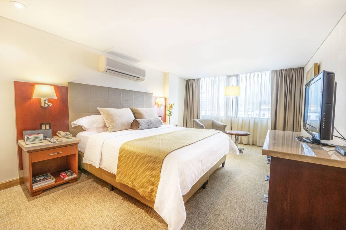 Una habitación de hotel con cama, mesitas de noche, escritorio, televisión y aire acondicionado.