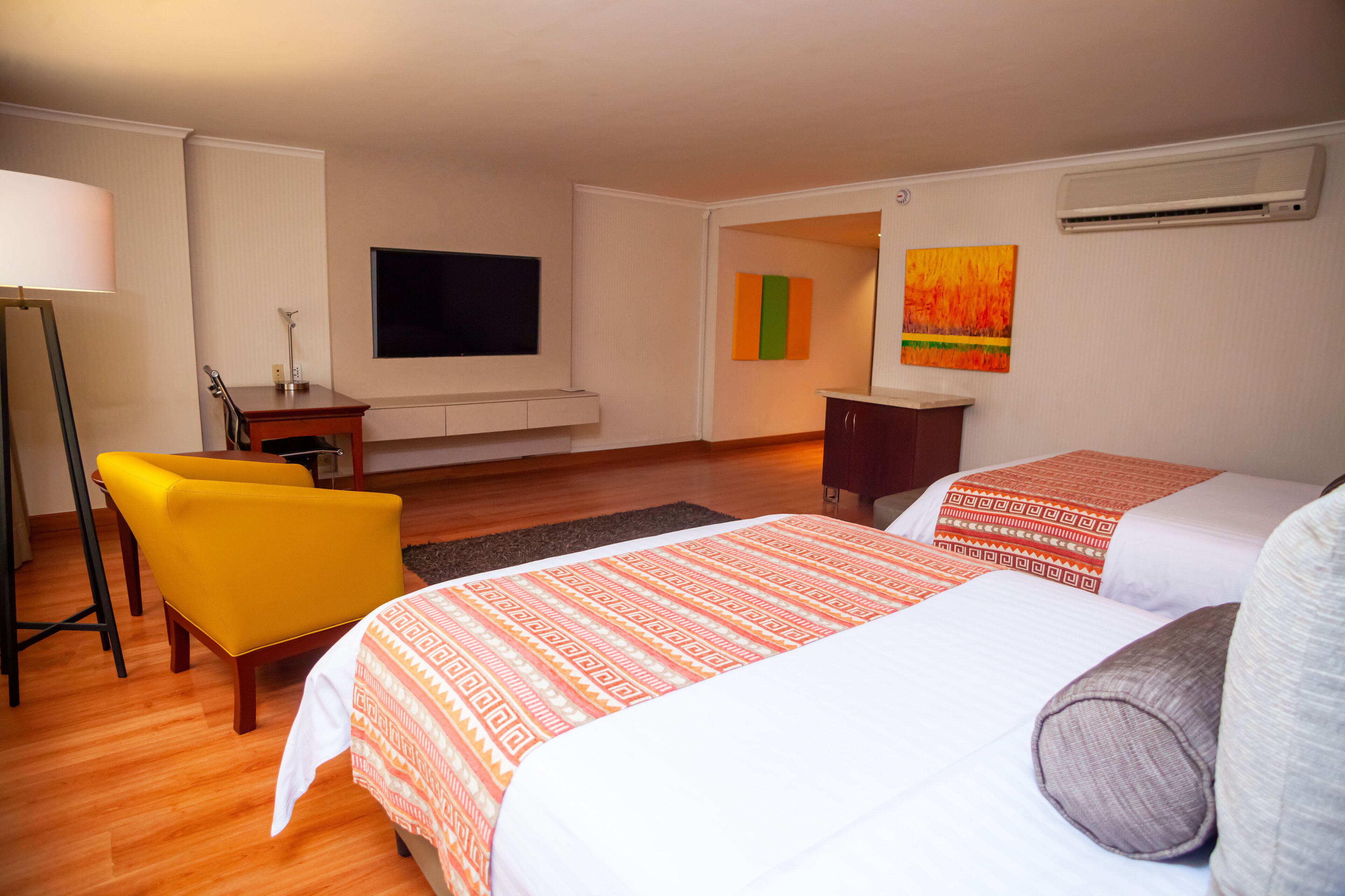Una habitación de hotel con dos camas, un escritorio, una silla, un televisor y obras de arte en las paredes.