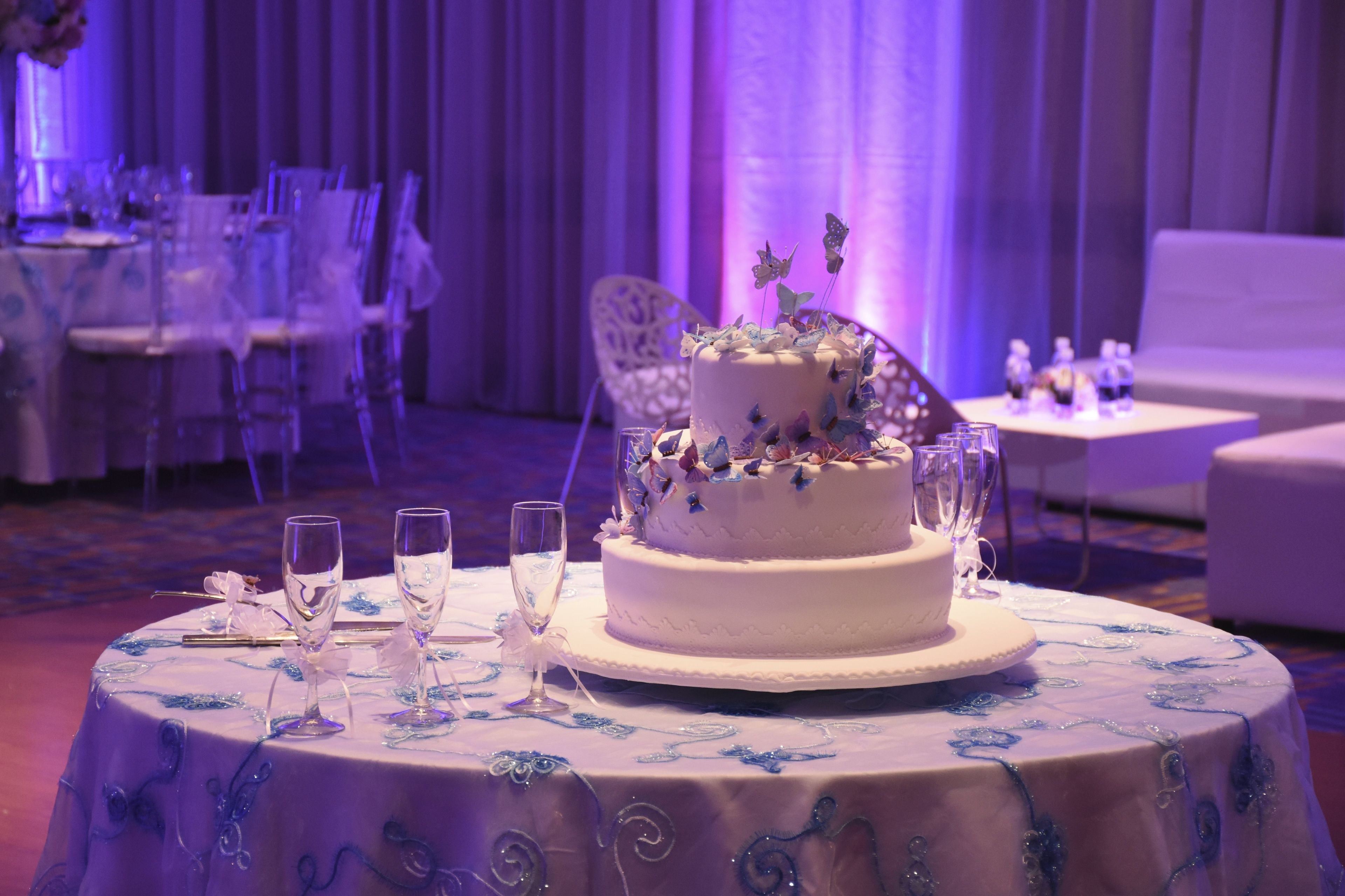 A round table with a white cake decorated with butterflies, surrounded by clear glasses and a white tablecloth with blue patterns.