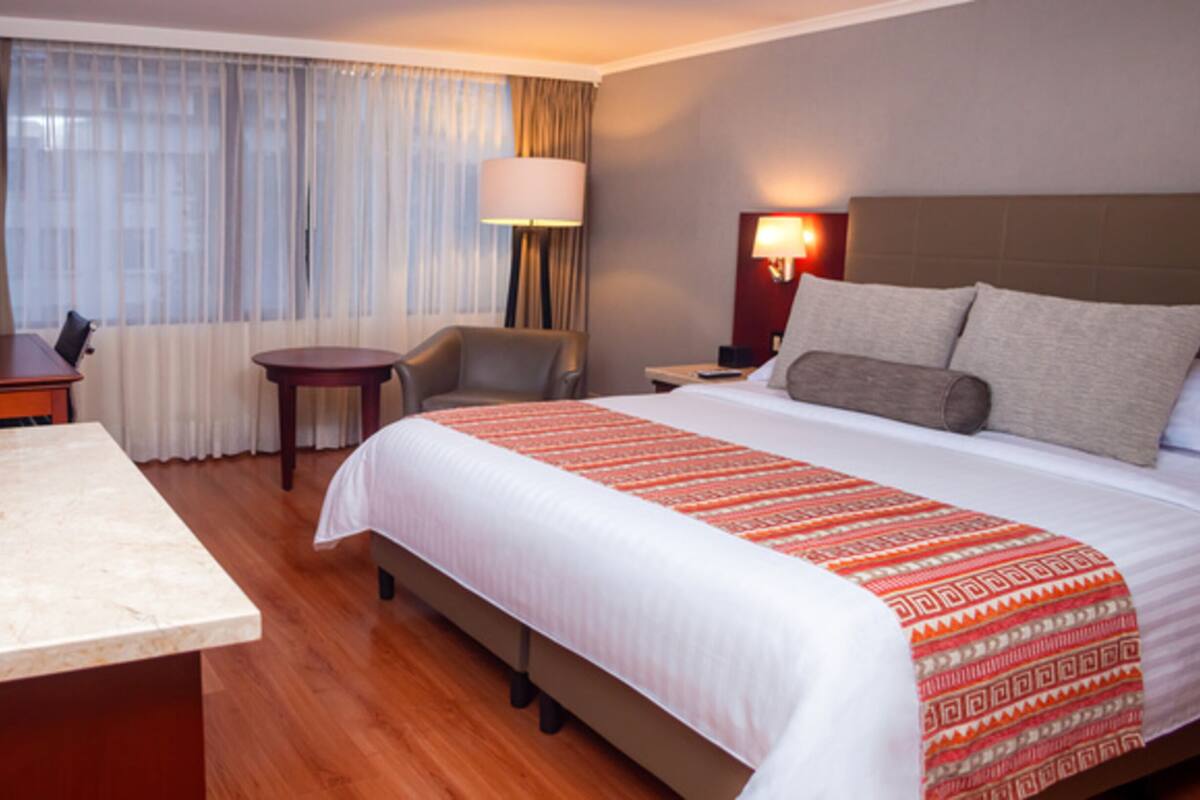 Habitación de hotel con una cama grande, un escritorio, una silla y una mesita de noche con lámpara.