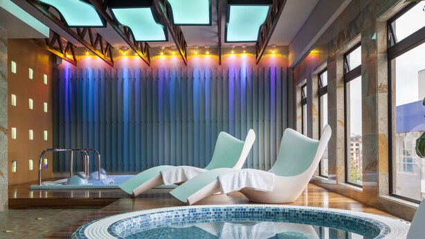 A modern indoor pool area with a circular hot tub, lounge chairs, and a large window providing a view of the city. - Bogota Plaza Hotel  (Bogotá)