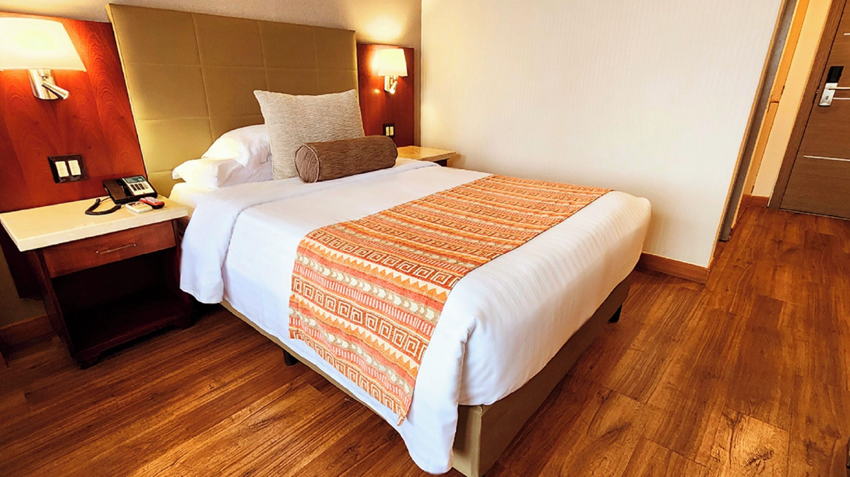 Quarto de hotel com uma cama grande, cabeceira de madeira, mesas de cabeceira com luminárias, telefone e piso de madeira.
