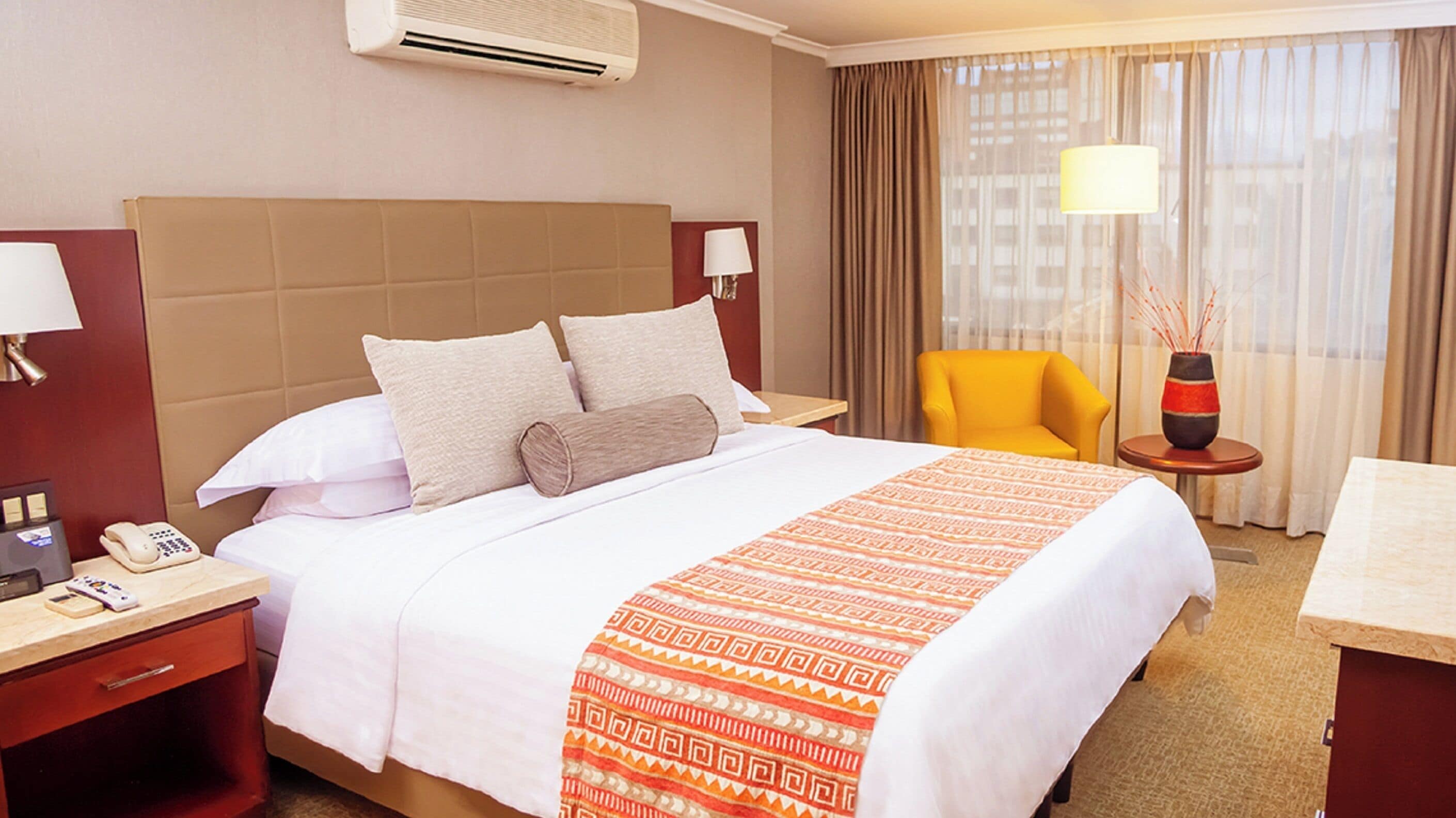 A hotel room with a large bed, a bedside table with a phone, a chair, a lamp, and a vase with decorative sticks.