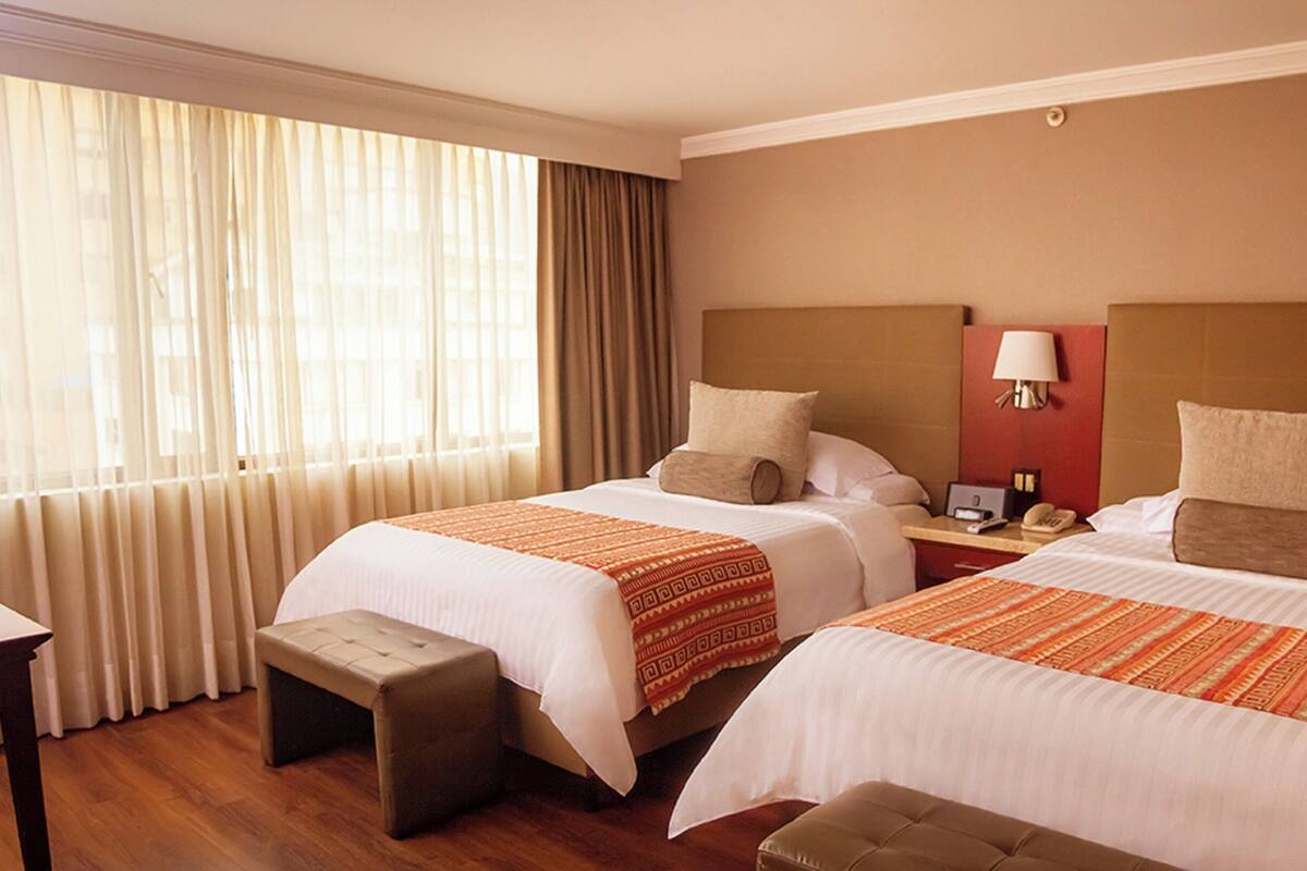 Habitación de hotel con dos camas, una mesita de noche, una lámpara y una ventana con cortinas.