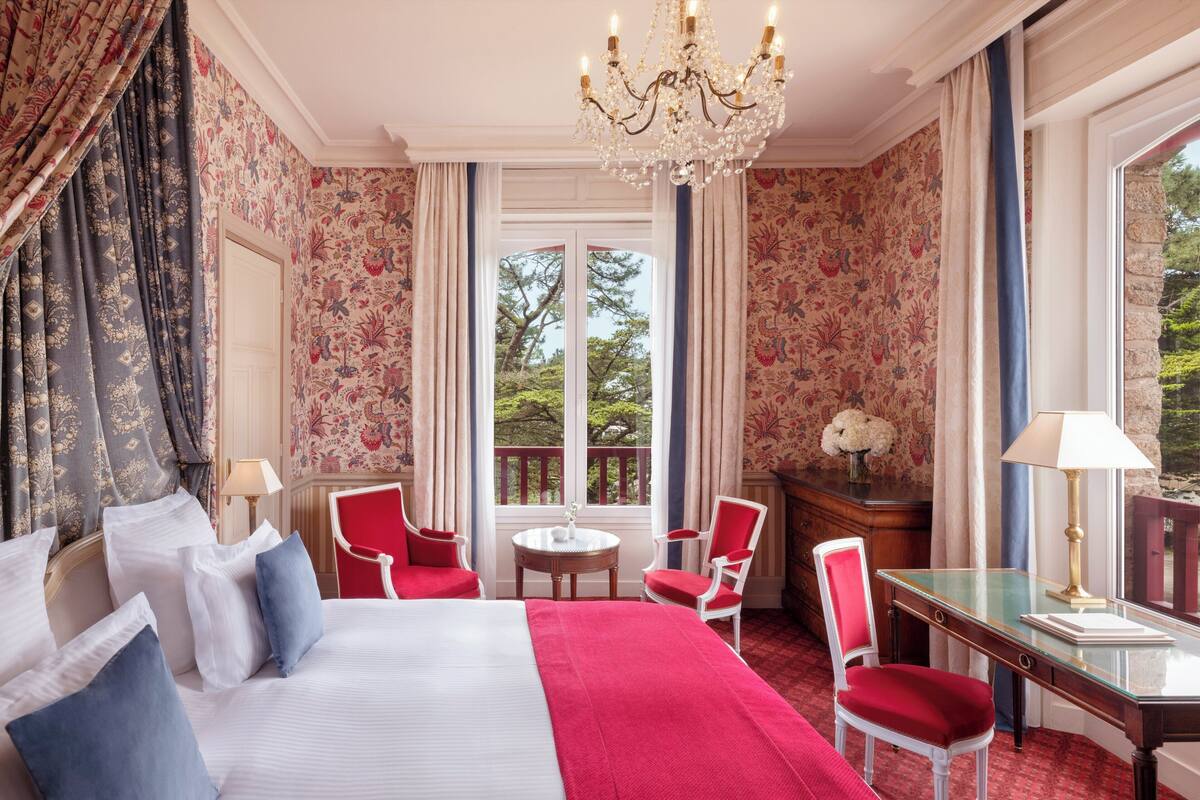 A hotel room with a large bed, red chairs, a desk, and a chandelier.