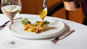 A dining table with a plate of food, a glass of white wine, and a person pouring sauce from a small pot.
