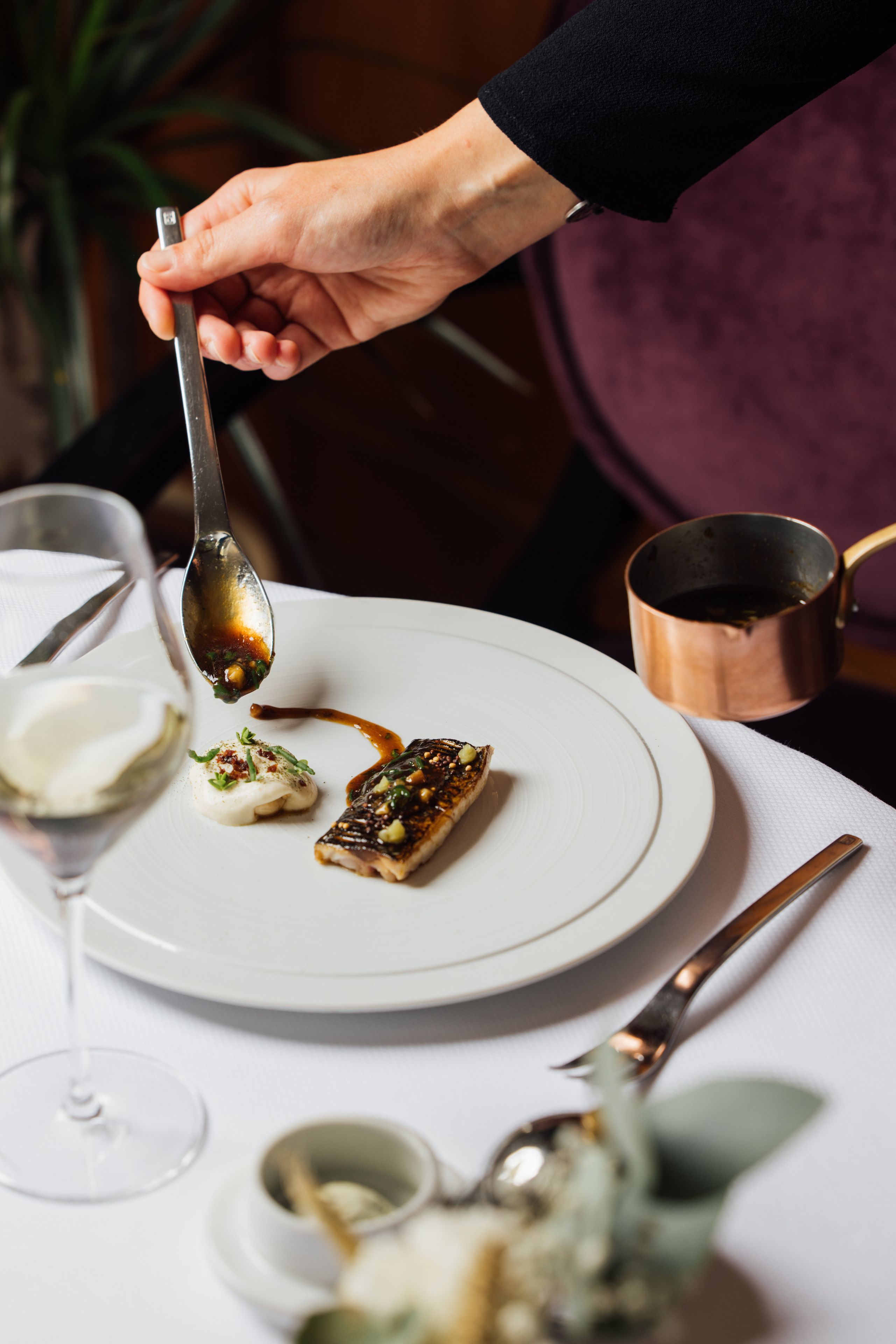 A dining table with a plate of food, a spoon, a glass of white wine, and a copper pot.