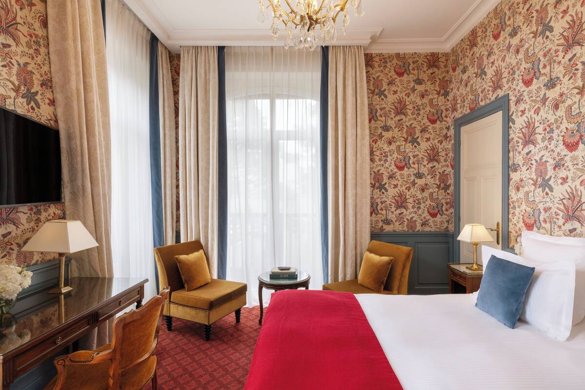 A hotel room with a large bed, two armchairs, a desk with a lamp, a small table, and a TV.