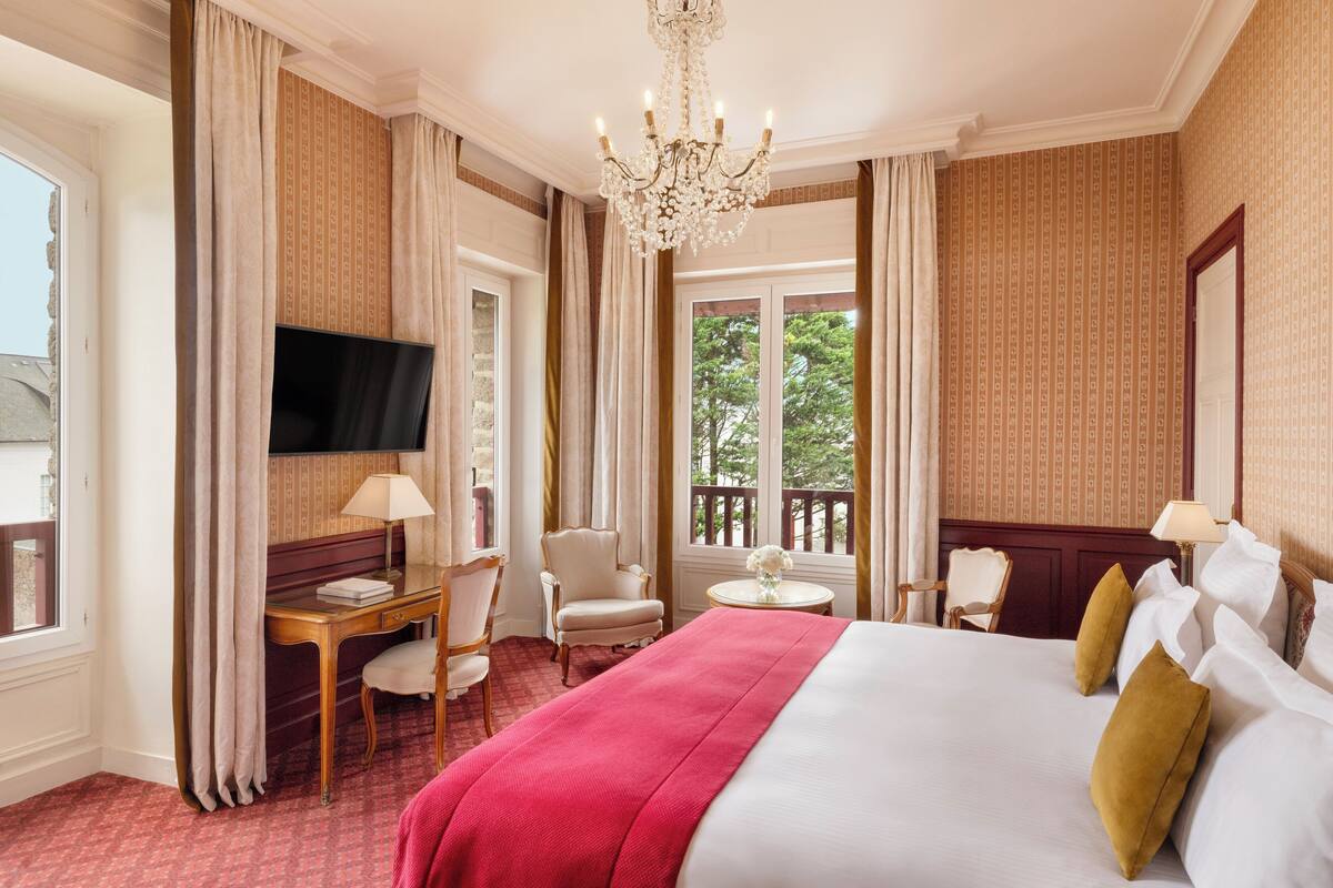 A hotel room with a large bed, a desk with a chair, a television, and a balcony with a view of trees.