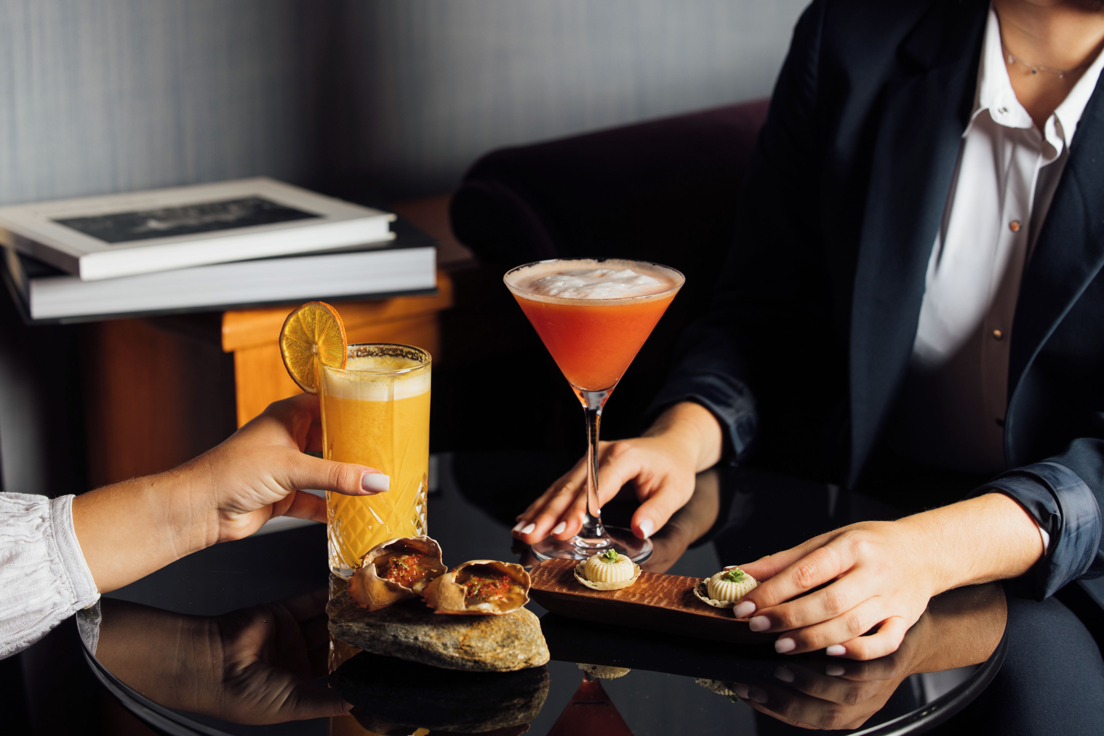 A person holding a glass of orange juice and a martini glass with a cocktail on a table.