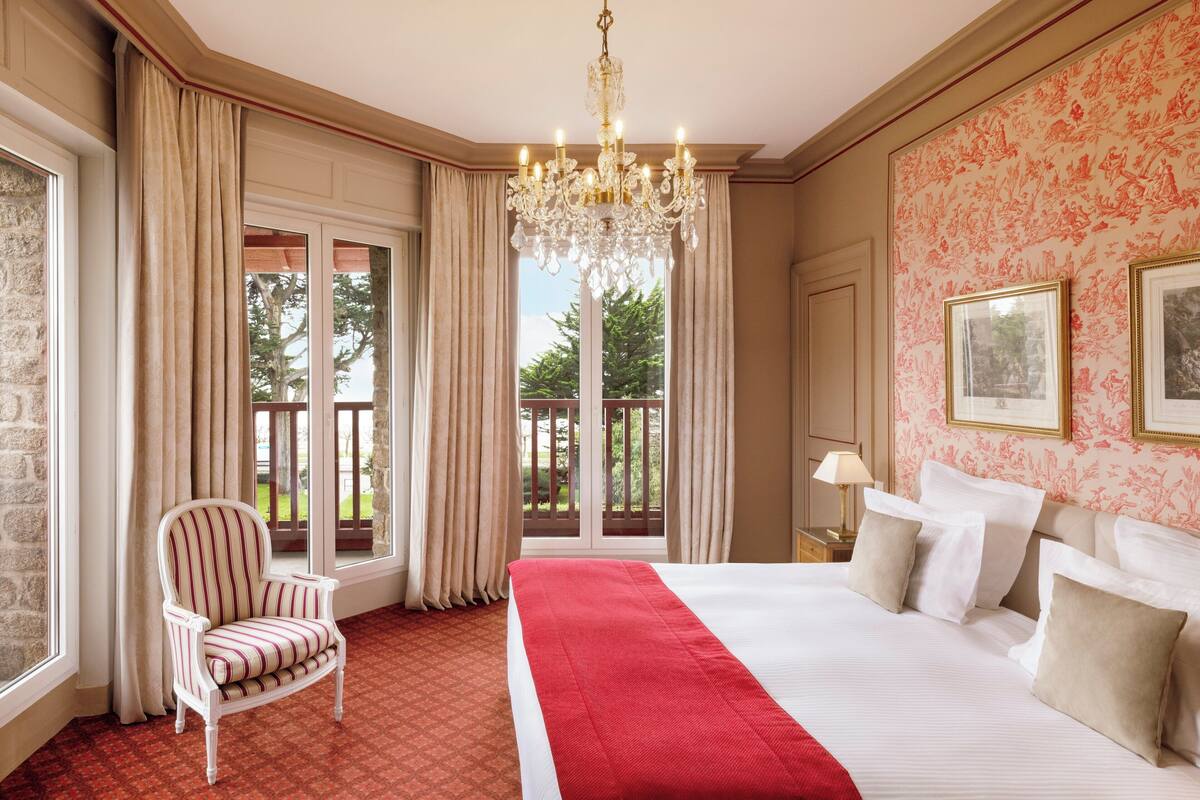 A hotel room with a large bed, a chandelier, a chair, and a balcony with curtains.