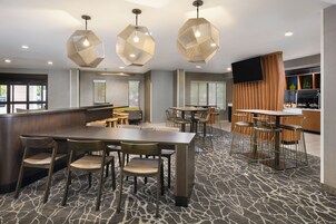 Un moderno vestíbulo de hotel con una zona de bar central, varios arreglos de asientos e iluminación colgante geométrica.