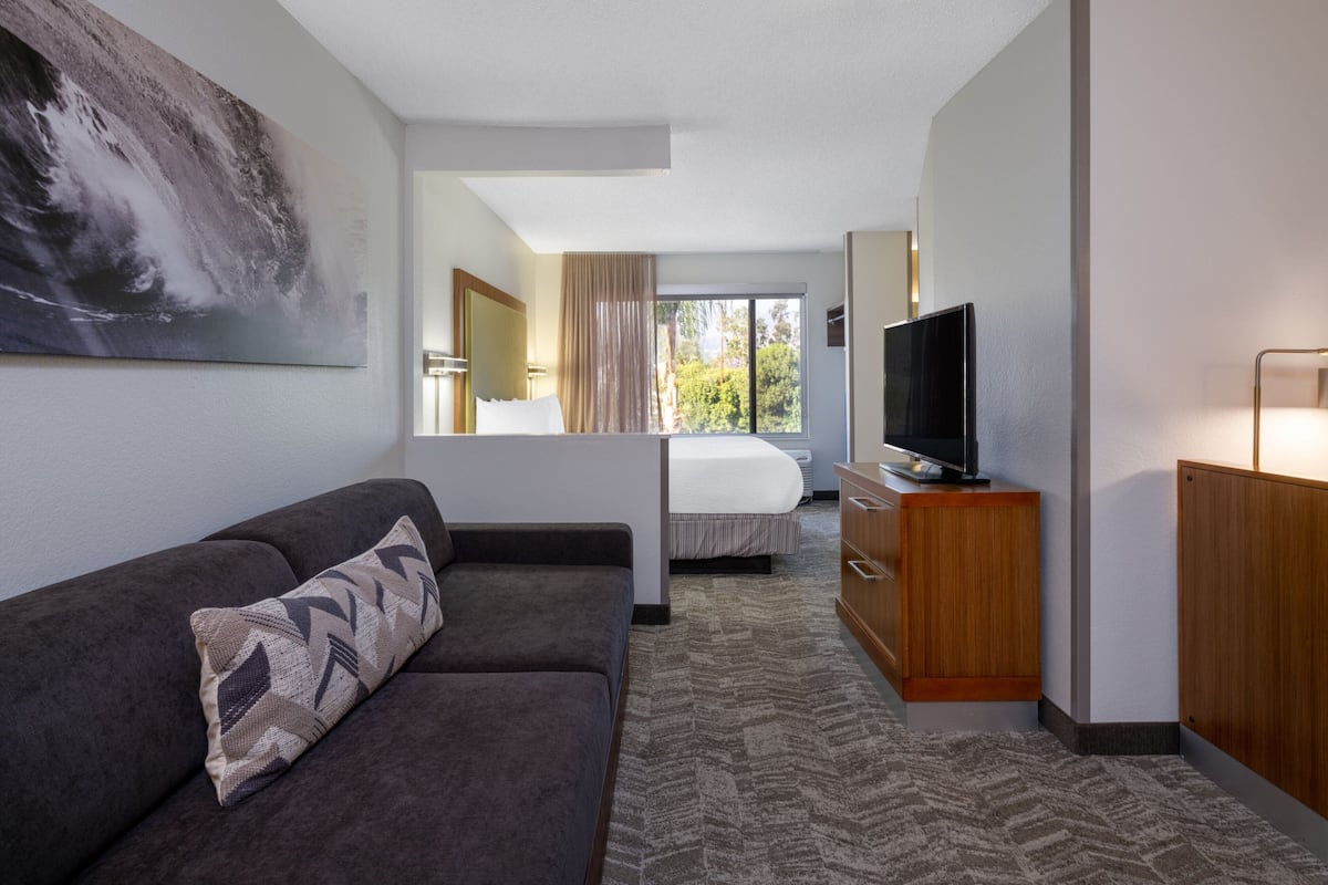 A hotel room with a sofa, a TV on a wooden stand, a bed with a window view, and a large painting on the wall.