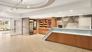 Un hall d’hôtel moderne avec un comptoir d’accueil, des étagères en bois où sont disposés divers articles de confort, un réfrigérateur et une entrée avec une porte en verre.