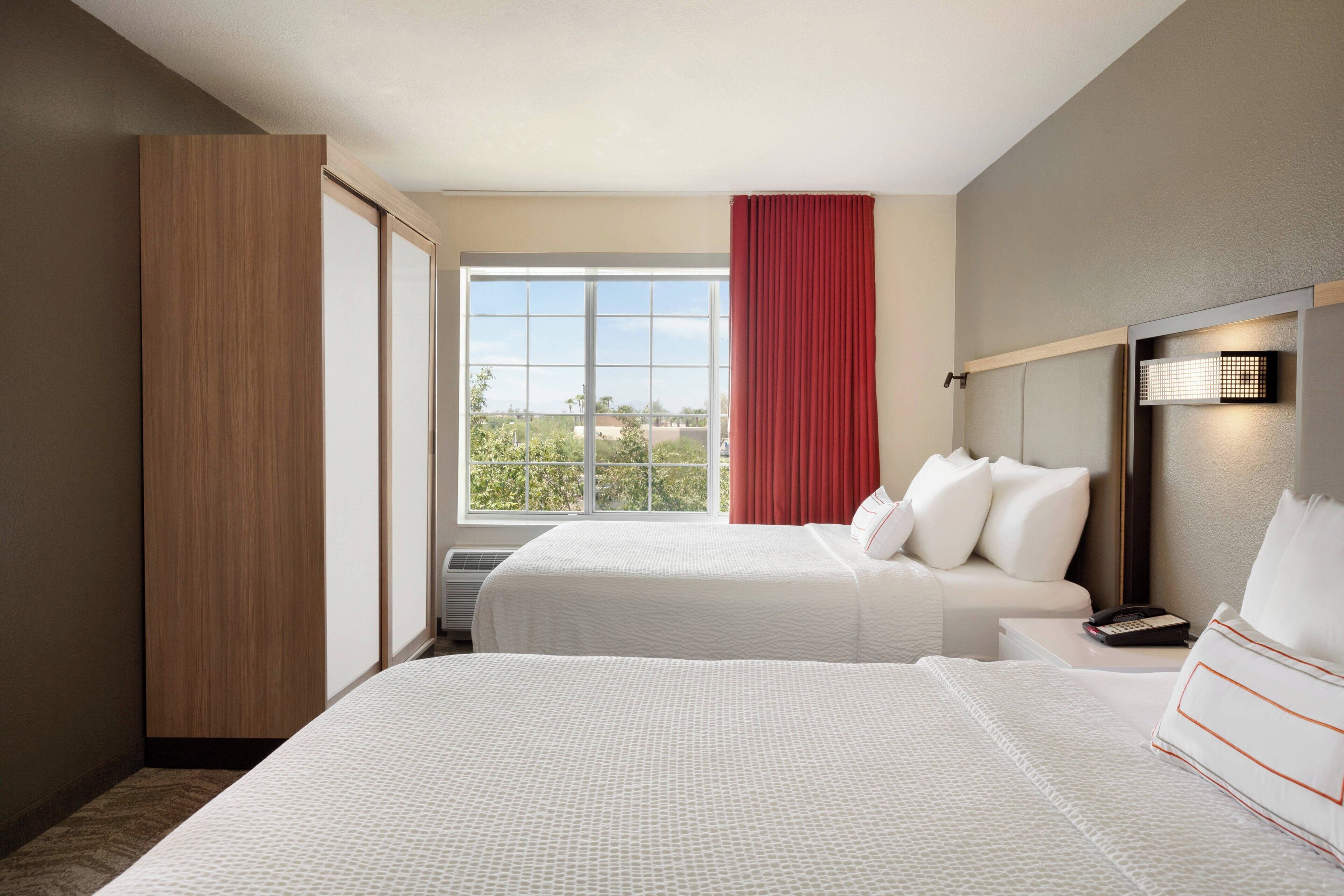 A hotel room with two beds, a large window, a red curtain, and a bedside table with a phone.