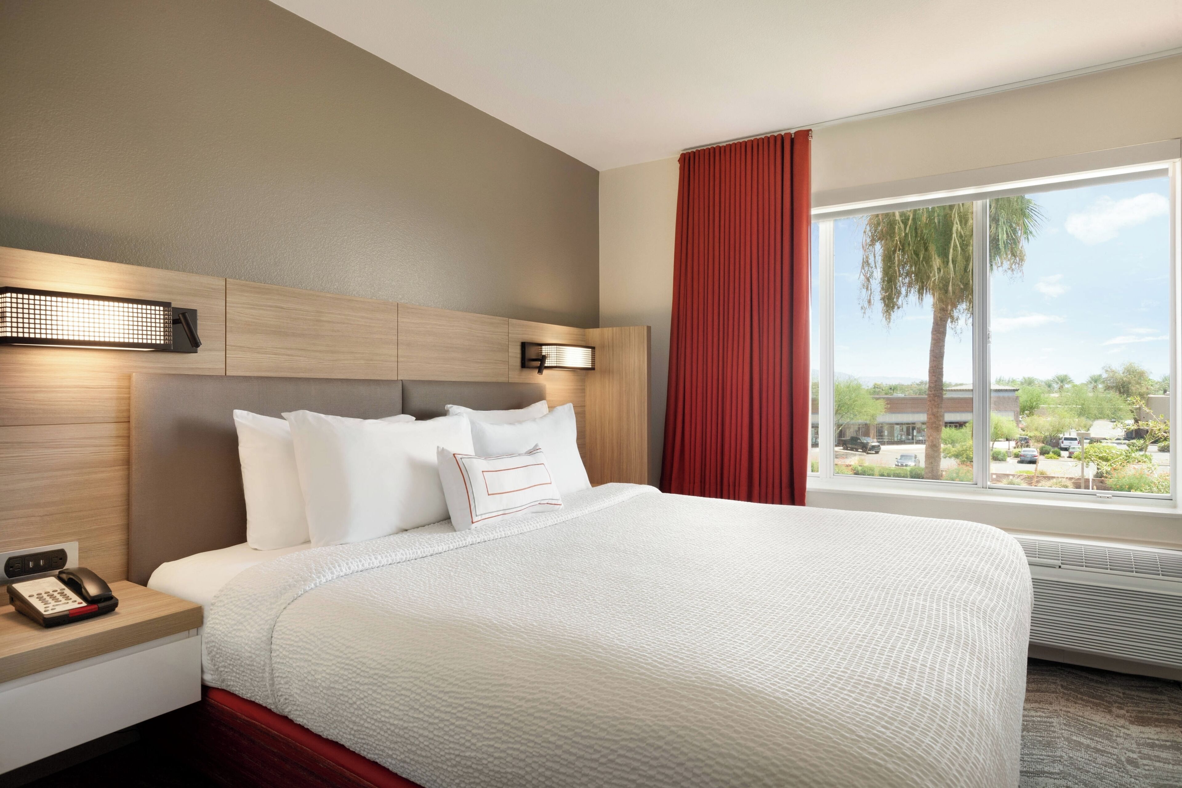 A hotel room with a large bed, a bedside table with a phone, and a window with a view of the outside.