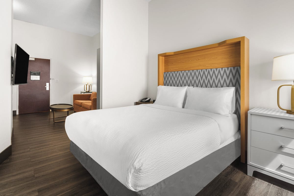 A modern hotel room with a large bed, a wooden headboard, a bedside table with a lamp, a small round table, and a television mounted on the wall.