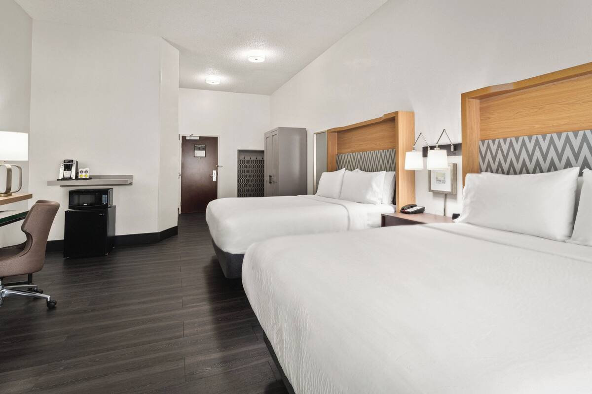 A hotel room with two beds, a desk, a chair, a wardrobe, and a small kitchenette.