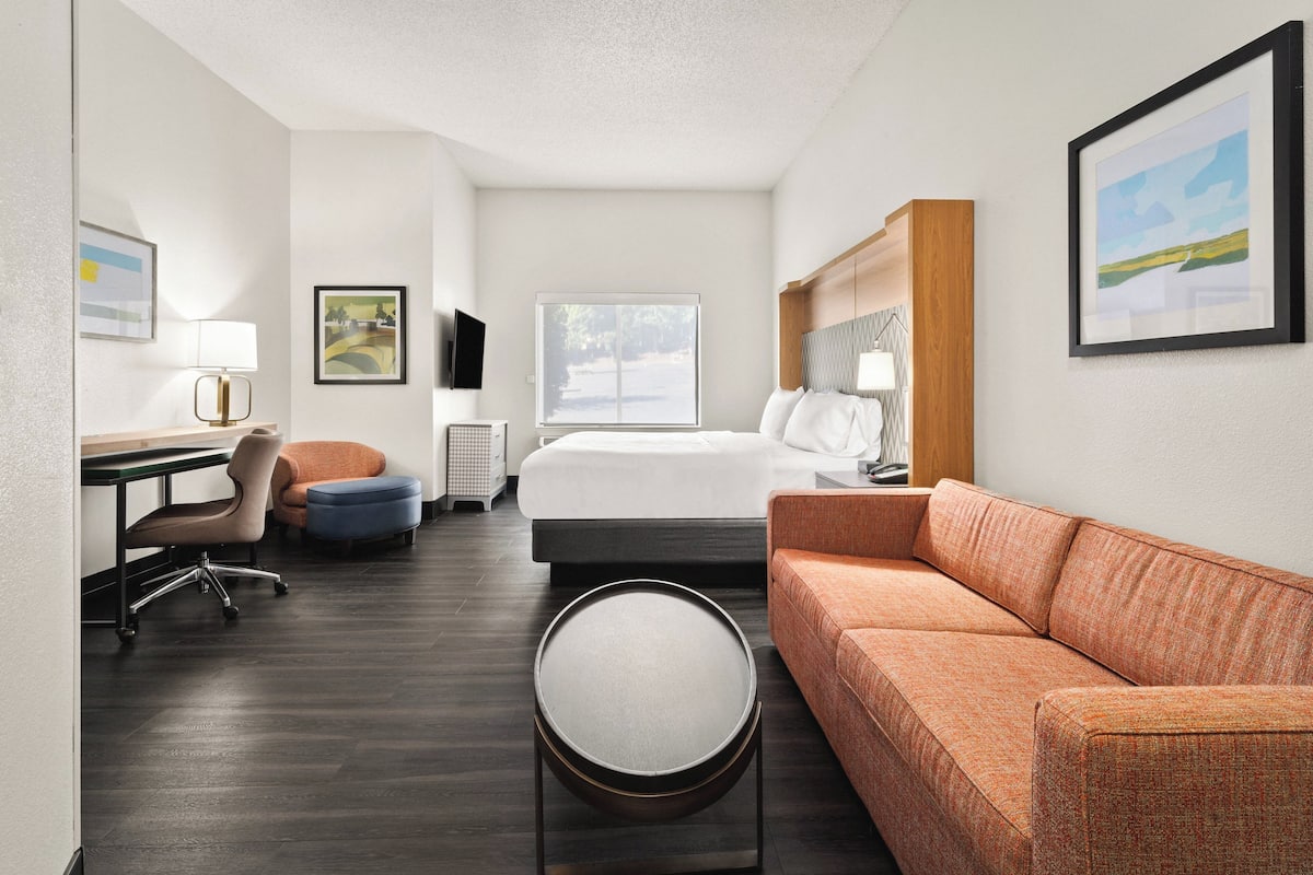 A hotel room with a bed, a sofa, a desk with a chair, a round table, and a TV on the wall.