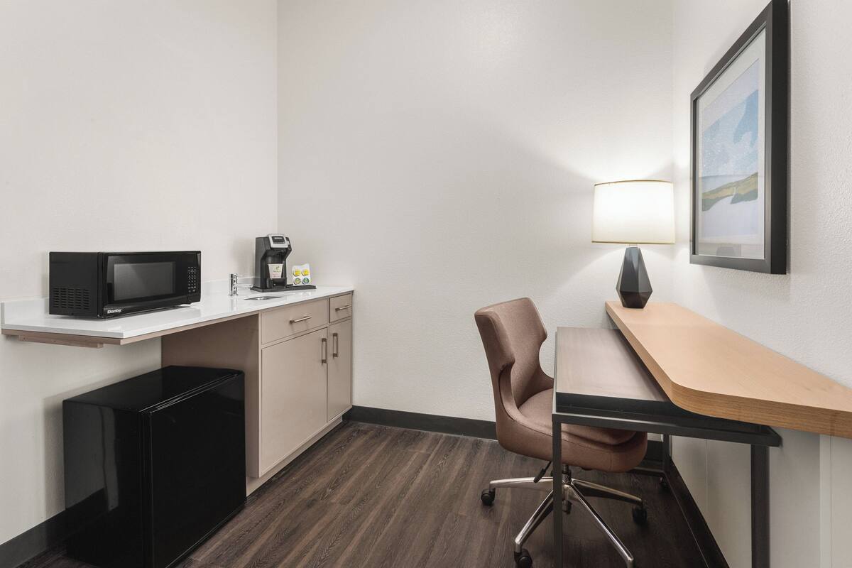 A compact hotel room with a kitchenette, a desk, a chair, a lamp, and a framed picture on the wall.