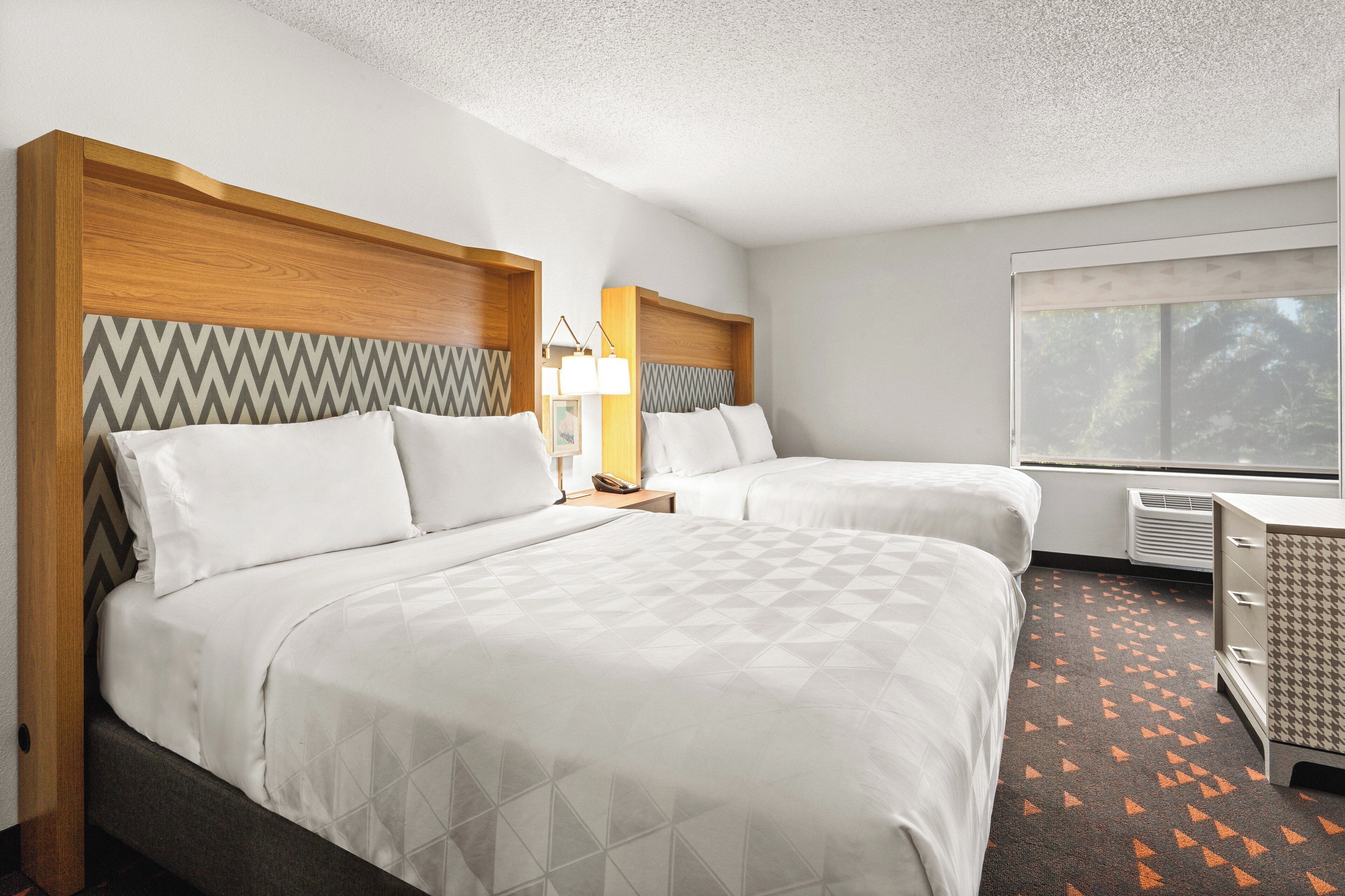 Habitación de hotel con dos camas, una mesita de noche con lámpara, cómoda y ventana con vista a árboles.